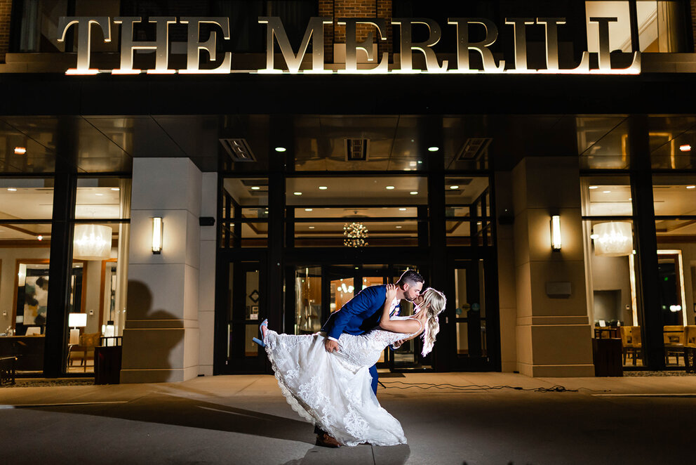 Man holding and dipping his bride in a wedding dress in front of The Merrill Hotel