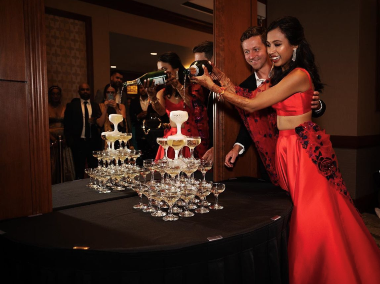 A bride and groom pouring champagne over a tower of champagne glasses with a mirror in the background reflecting their wedding guests